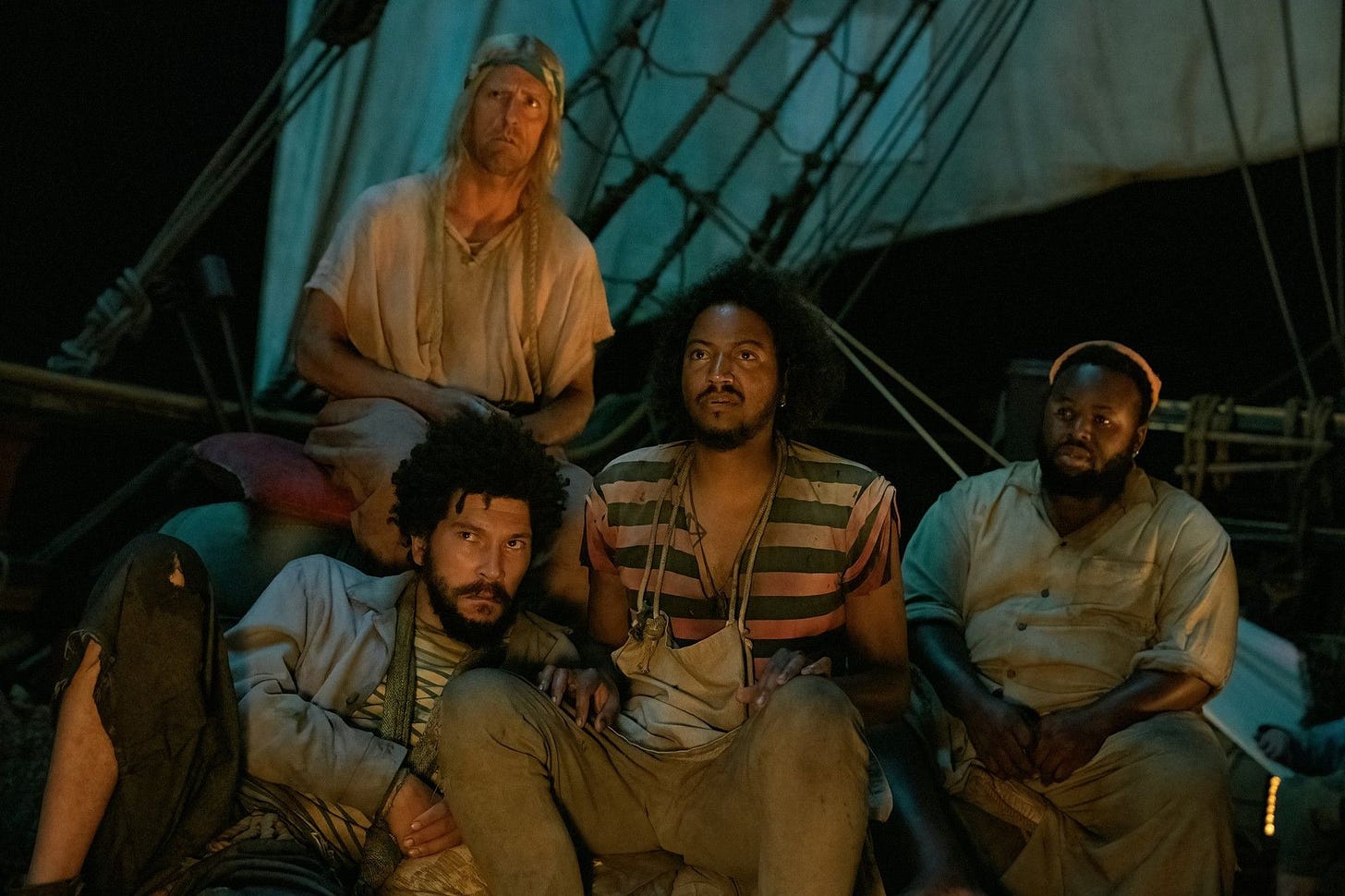 four pirates, a tall white man with blond shoulder length hair, a black man with curly hair and a beard, a black man and with messy curly hair and a beard wearing an apron and a striped shirt and a black man with a beard, an earring and an orange beanie all look with confusion at something off-screen