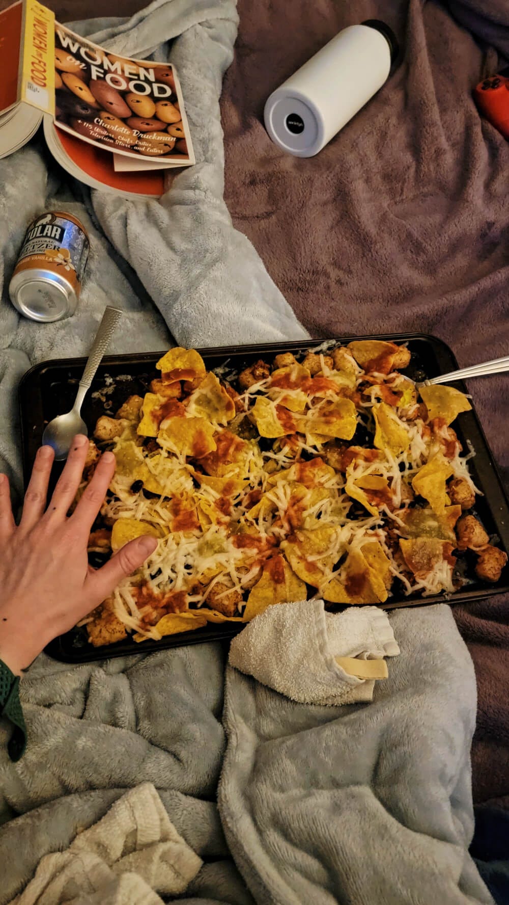 The main attraction of this dimly lit image is a dark metal sheet tray overflowing with a pile of tater tots, corn tortilla chips, tomato and tomatillo salsas, vegan mozzarella cheese, rested on top of two cozy blankets. Posted next to Grace’s small hand, for scale, taking up about 1/6 of the tray. Other items splayed across the blankets include: an orange vanilla Polar seltzer, the anthology, Women on Food, a water canteen, napkins, and the edge of a play station controller. 