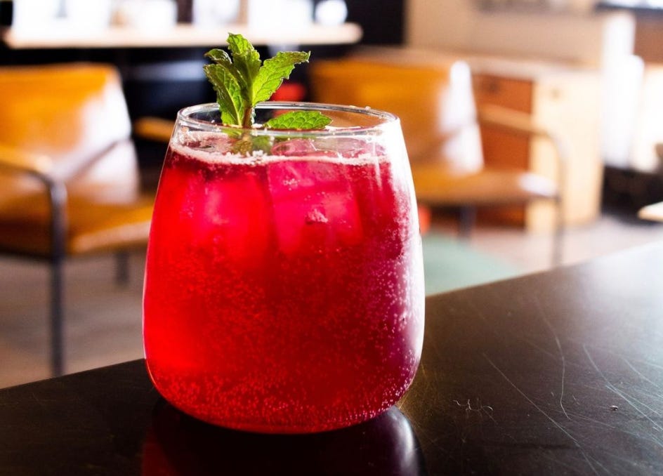 A close up of a red, fizzy drink in a flat bottomed juice glass with a mint leaf stick out. Labeled the, "blueberry mint sparkling lemonade."