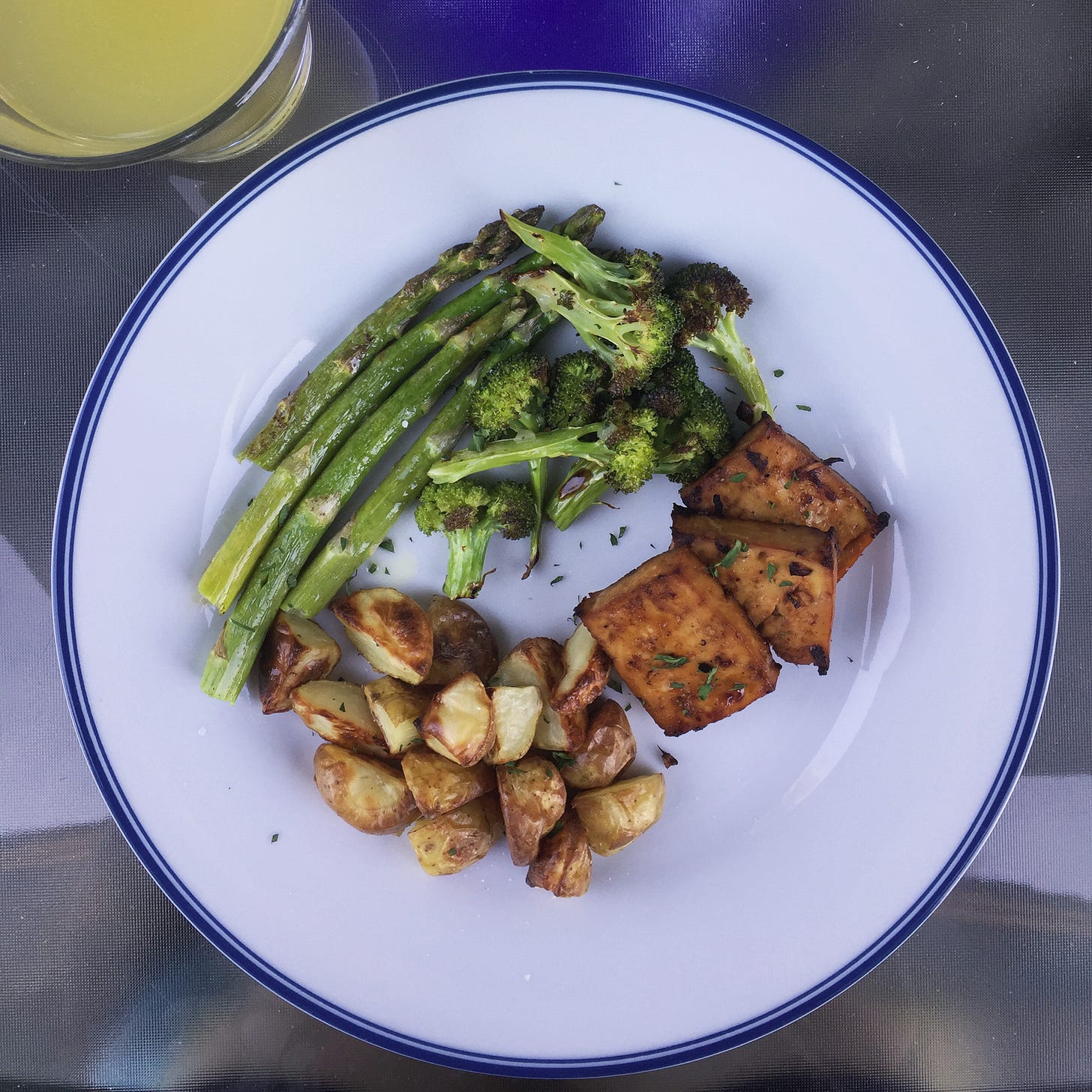 from above, a white plate with a blue rim holds small piles of roasted potatoes, broccoli, and asparagus. Three squares of marinated tofu sit to their right.