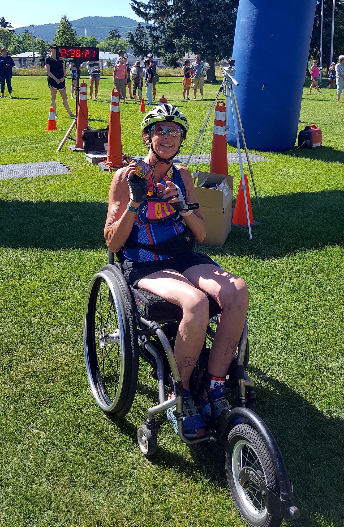 A woman in a white cycling helmet, cycling gloves, sunglasses, shorts and a sleeveless blue and pink athletic top sitting in a wheelchair holds up a finishers medal with a race finish line and people milling about behind her.