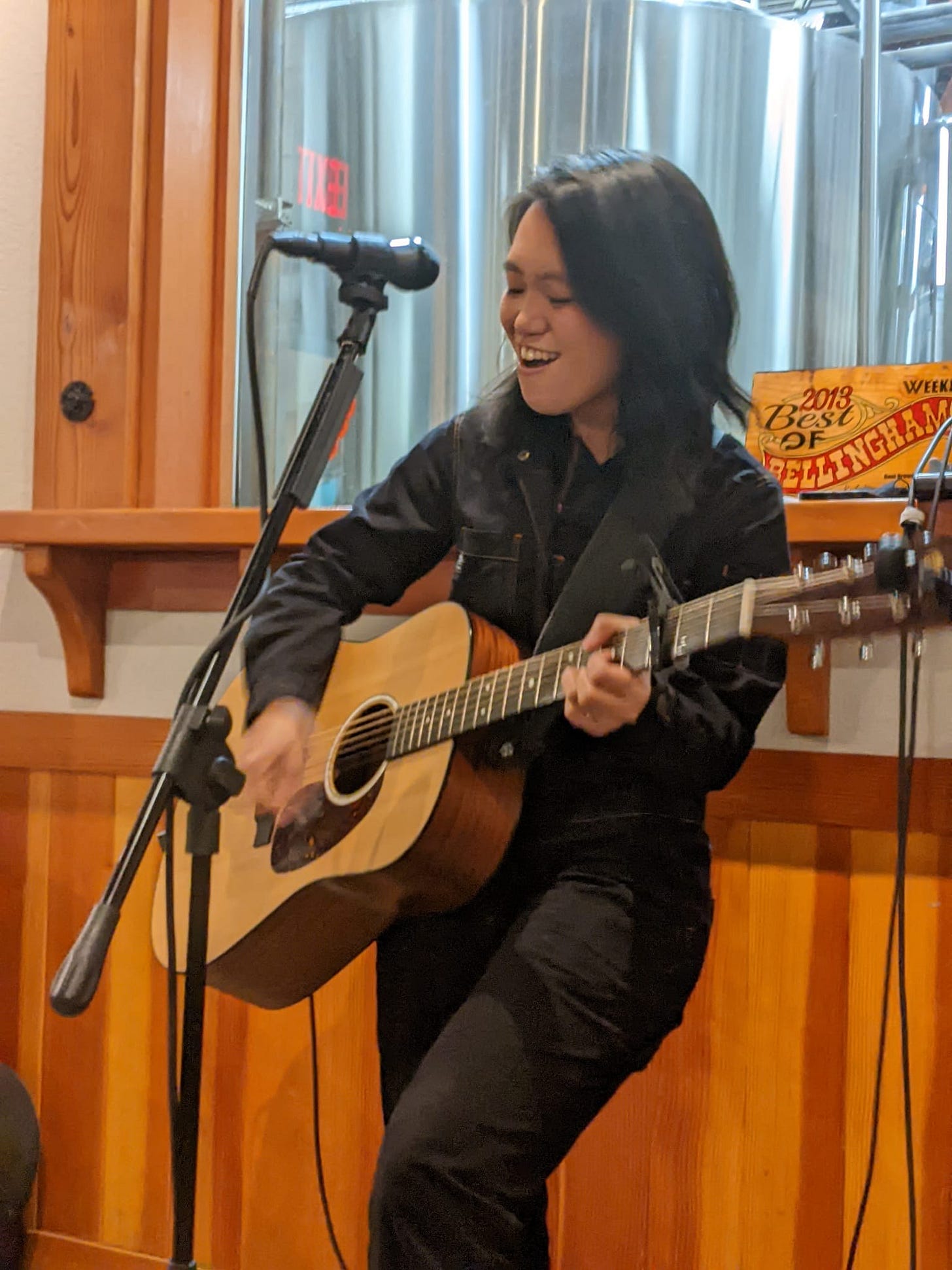 a singer with an acoustic guitar in front of some brewing equipment