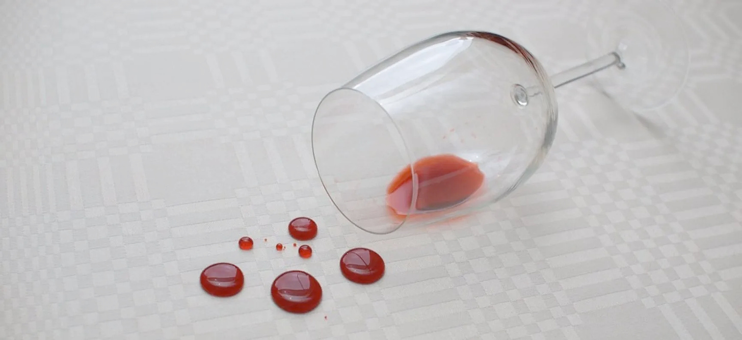 A near-empty wine glass on its side on a white tablecloth, with several spilled drops of wine
