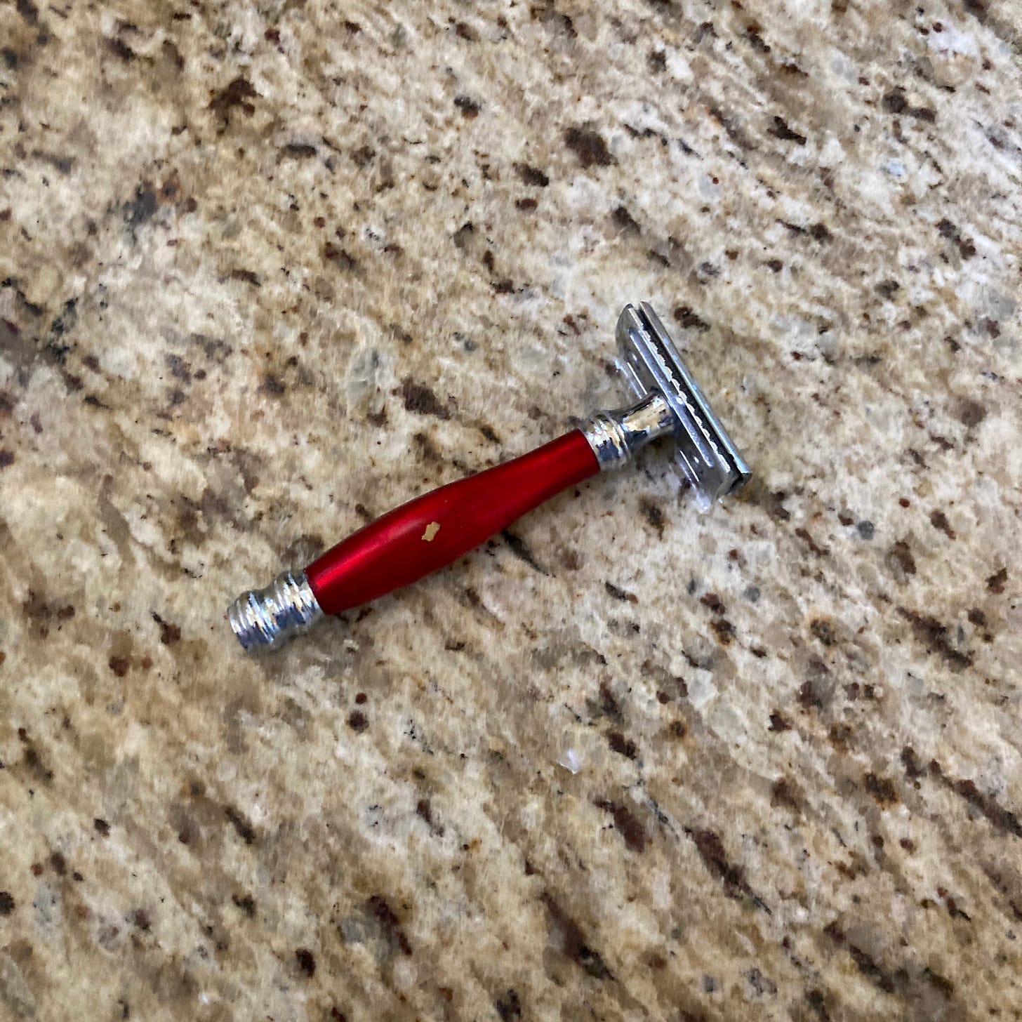 Visual description: A metal safety razor with a silver end, red handle, and silver top rests on a gray and brown granite countertop.