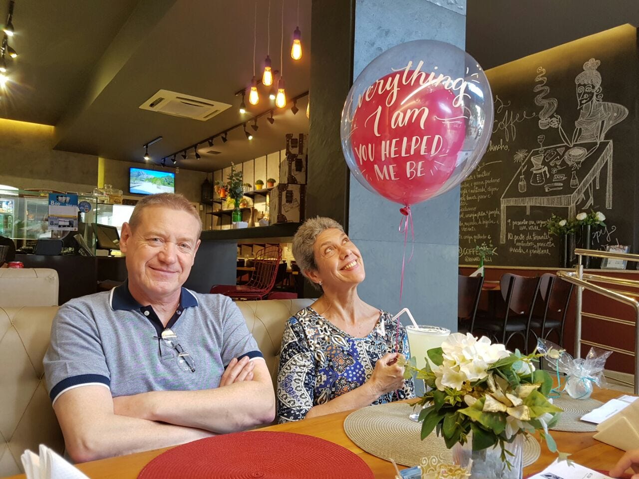 A white woman with white hair and a white man with blond hair, both in their 60s, sit in a restaurant booth. The woman holds a ballon.