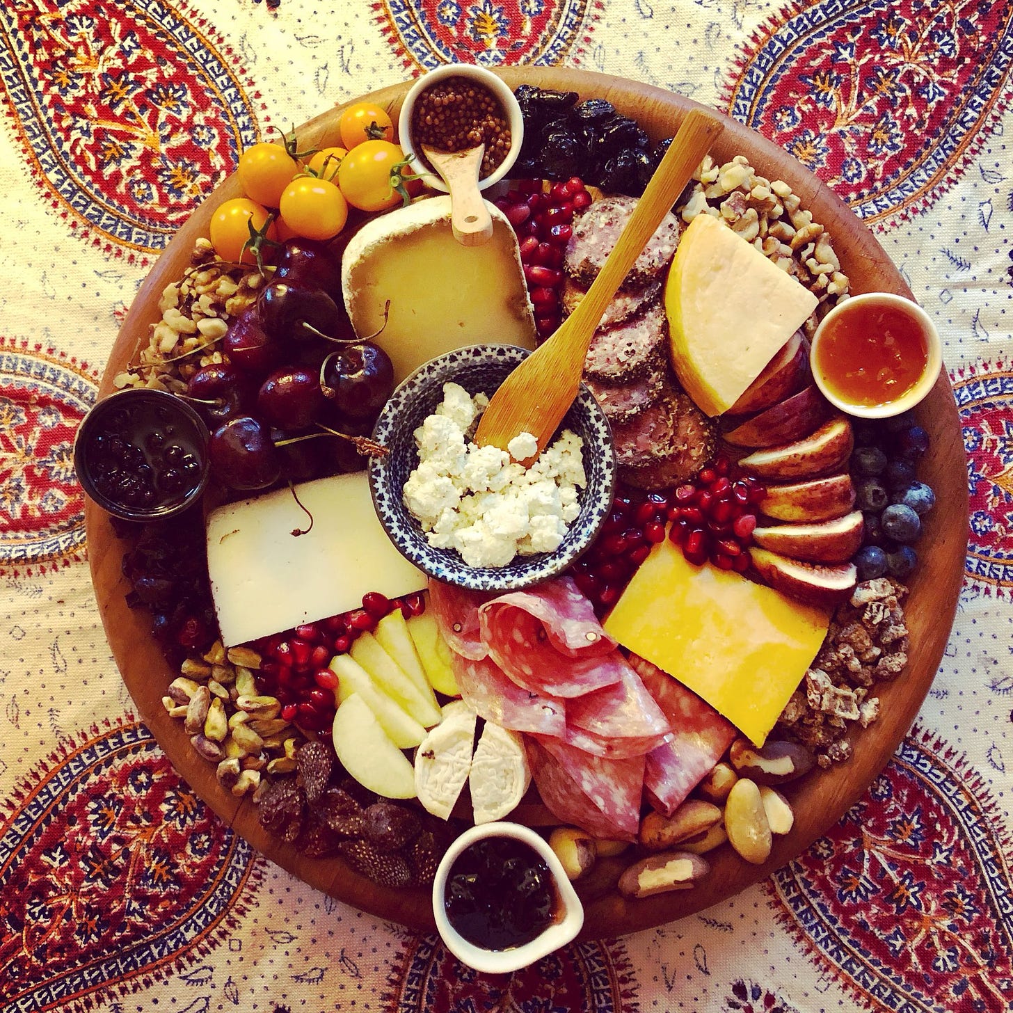a large round cheese board with cheeses, fruits, meats, and nuts on a persian tablecloth