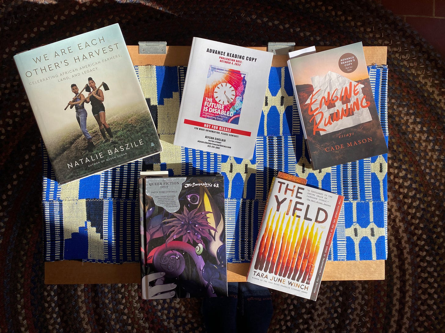 Six books arranged face-up on a coffee table covered in a blue cloth: We Are Each Other’s Harvest, The Future is Disabled, Engine Running, The McSweeney’s Queer Fiction Issue, and The Yield.