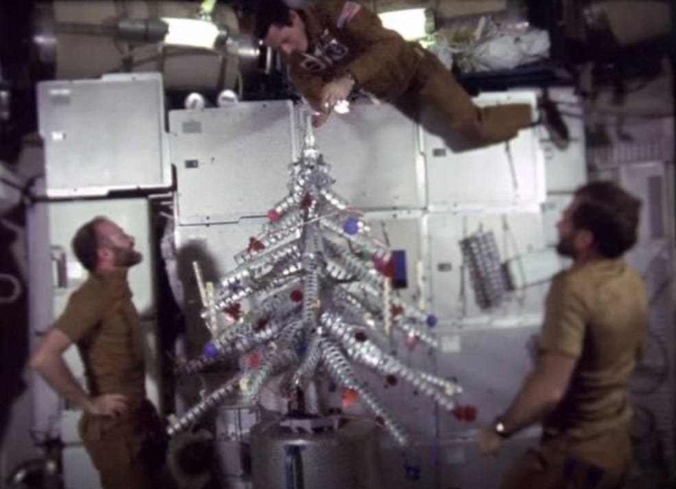 Color photograph of three white male astronauts with brown hair floating inside the Skylab 4 station around a shiny metal Christmas tree, bearing a few colored circle as decorations. One astronaut floats directly above the tree, adding a light to its top.
