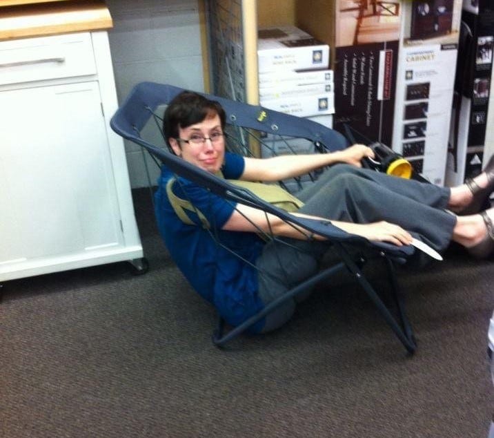 A white woman with short brown hair and glasses, wearing a blue short-sleeved shirt and gray trousers, has fallen backside first through gaps in the cords of a bungee chair.