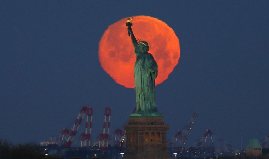 The moon is seen behind the Statue of Liberty.