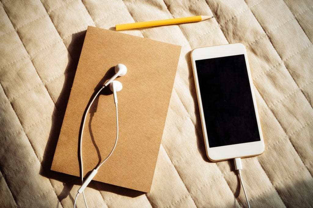 A light brown journal with a pencil above it and a white cellphone with white earbud headphones plugged into it lying next to the journal. These items are lying on the surface of a beige coverlet.