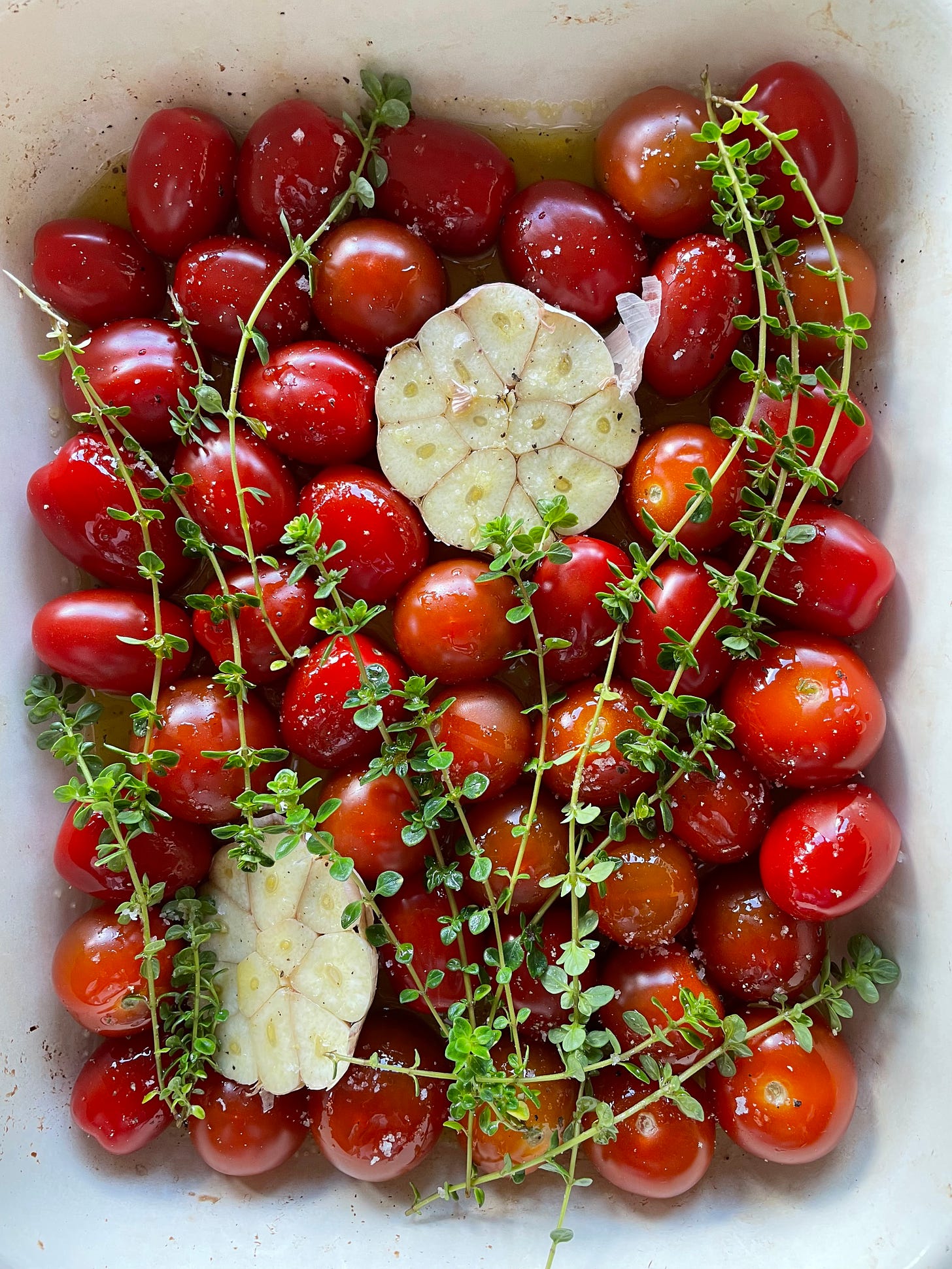 Roasting tray filled with cherry tomatoes, thyme, a halved head of garlic and plenty of olive oil.
