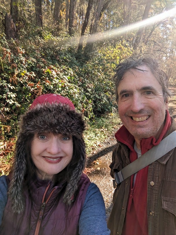 Blue-eyed woman in faux-fur trimmed red hat that covers her ears next to smiling brown-haired man against a sunny backdrop of autumn trees.