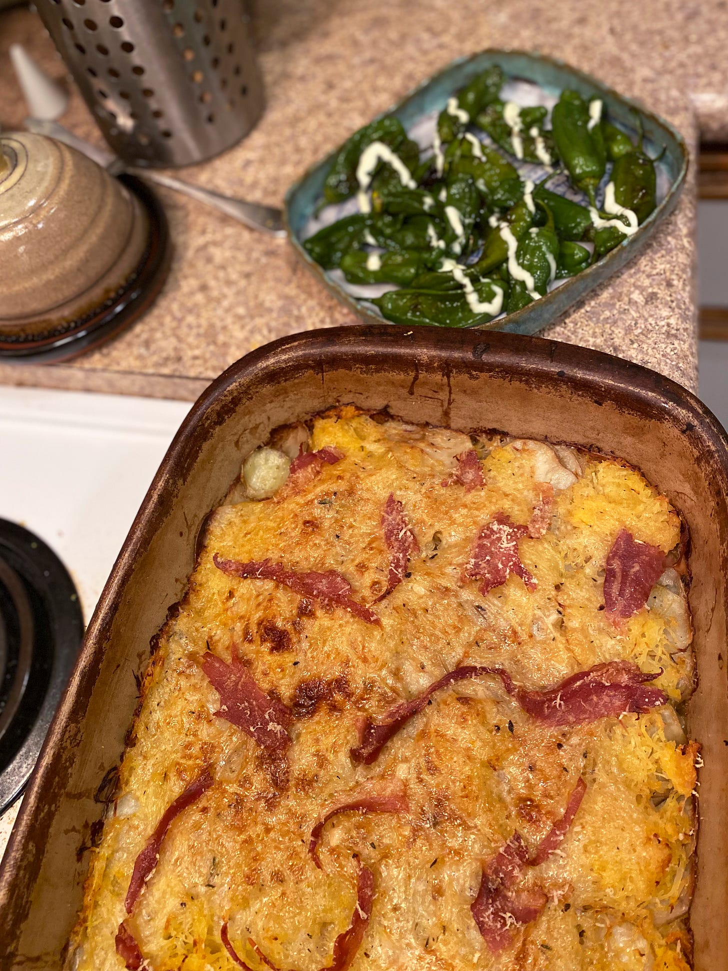 Resting on the stovetop is large rectangular stoneware dish of potato and spaghetti squash gratin, browned on top and crisp at the edges, with torn pieces of prosciutto peeking out from under the cheese. Behind it on the counter is a blue ceramic dish of blistered shishito peppers, with a drizzle of mayo overtop of them.