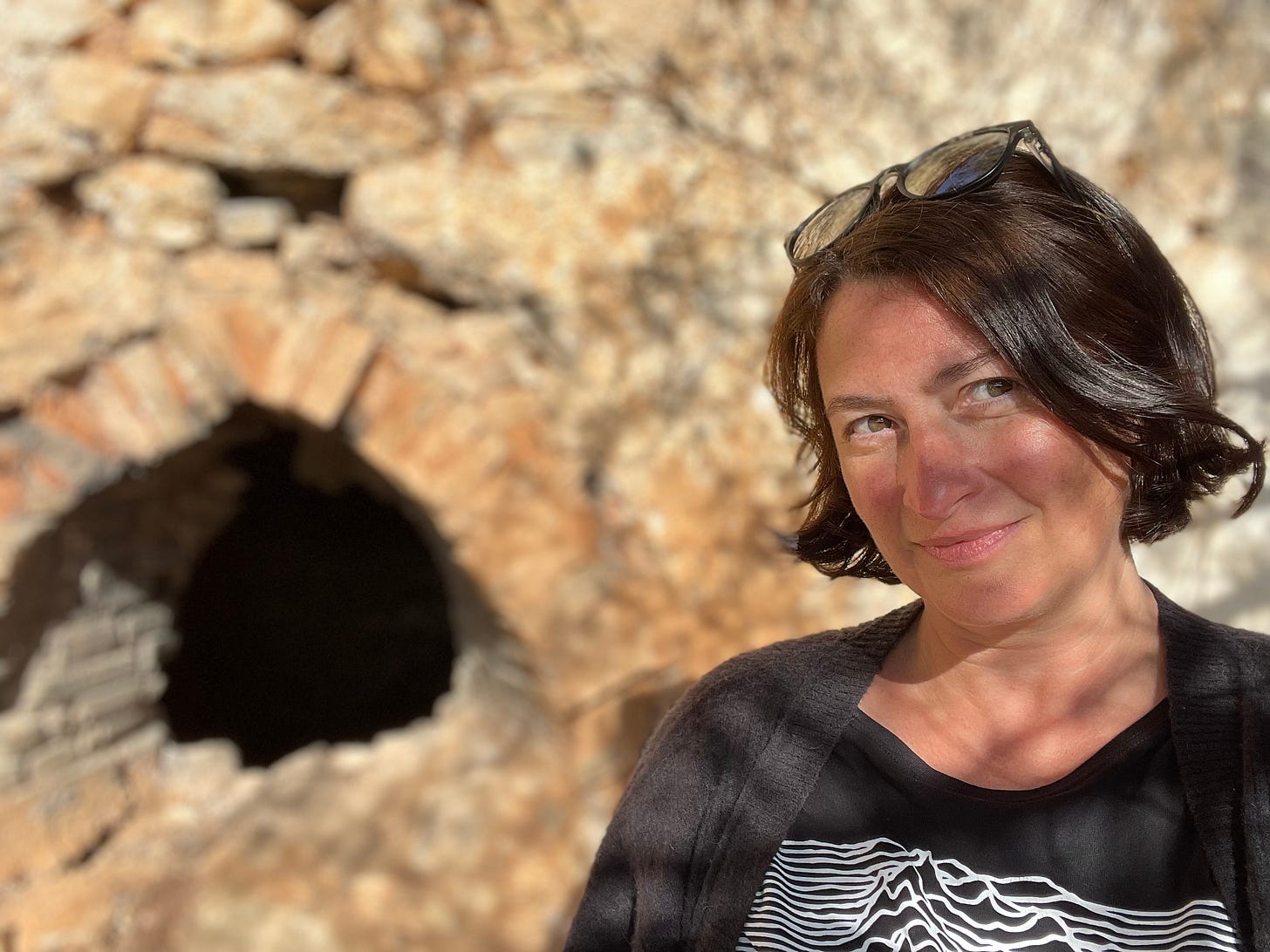 Colour photo of Sarah Farley standing in front of an old stone house, smiling and squinting at the sun