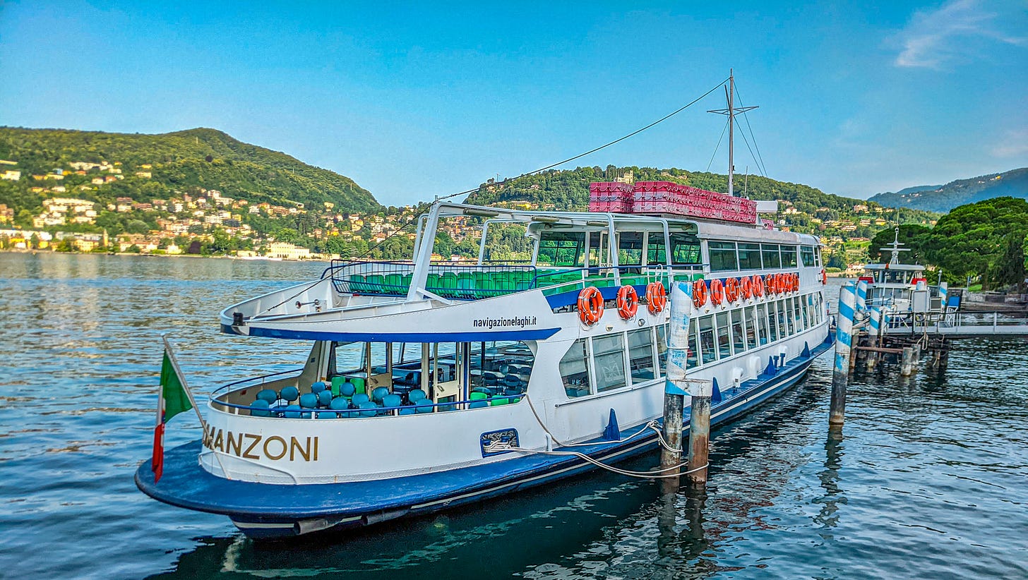 A large white and blue, two deck ferry sitting at a pier on Lake Como on a beautiful morning. 