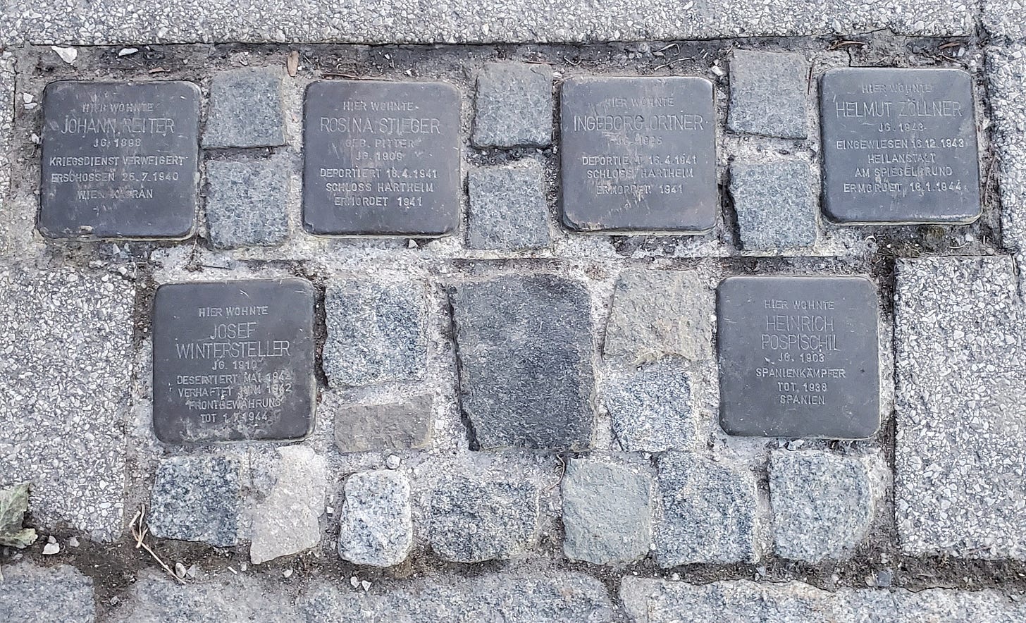 A group of six stumbling stones, with a seventh perhaps missing or destroyed, set in the cobblestone pavement. 