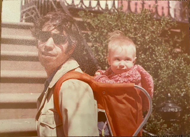 Dark-haired man with sunglasses wears a blond baby in a back carrier