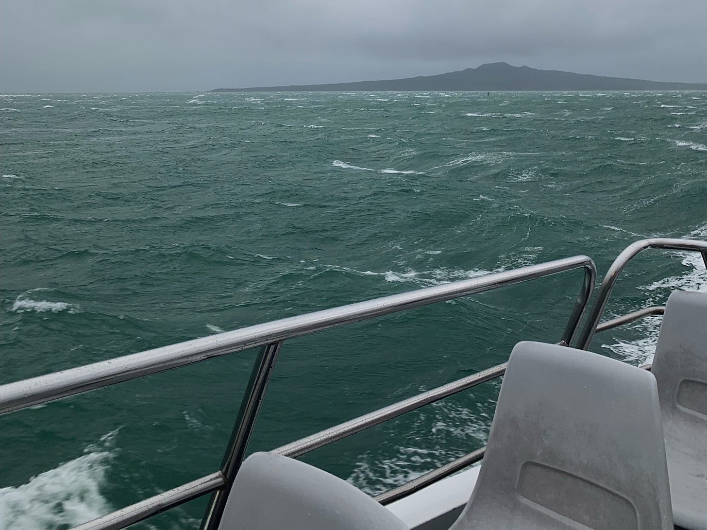 A wind-rucked sea carries a ferry across a harbour. The POV is from the top deck of a small ferry, with grey plastic seats in the foreground. In the mid-ground in the teal stromy sea and in the background is Rangitoto island, a dormant volcano against a dark grey sky
