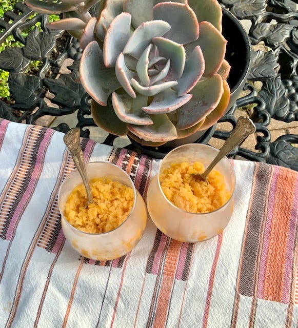 Two stemless wine glasses are filled with peach granita. They are laying on a striped towel. Each one has a spoon stuck upright in it.
