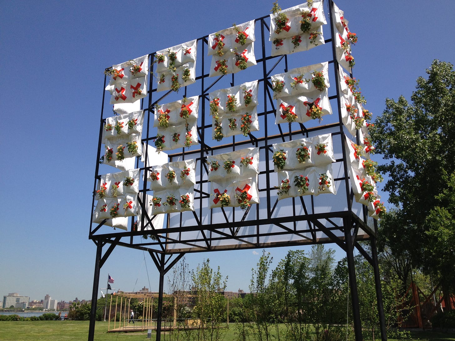 A large metal grid structure, as big as a highway billboard, has fabric bags hanging on three rows, with holes for plants to grow from. Each white bag is marked with a red "x" as part of Jeremijenko's "environmental health clinic."