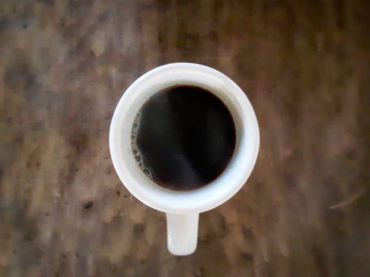 A top view of a ceramic coffee mug filled with black coffee, centered in the image with the handle pointing down and a blurred background of a dark wooden desktop.