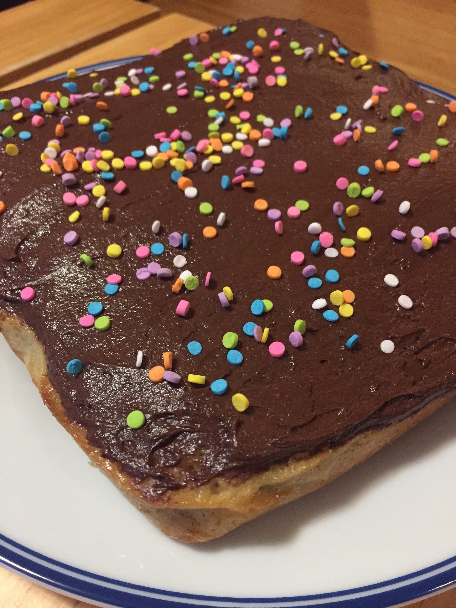 On a white plate, a square yellow cake with a layer of glossy chocolate icing. Large circular sprinkles in bright colours are scattered across the top.