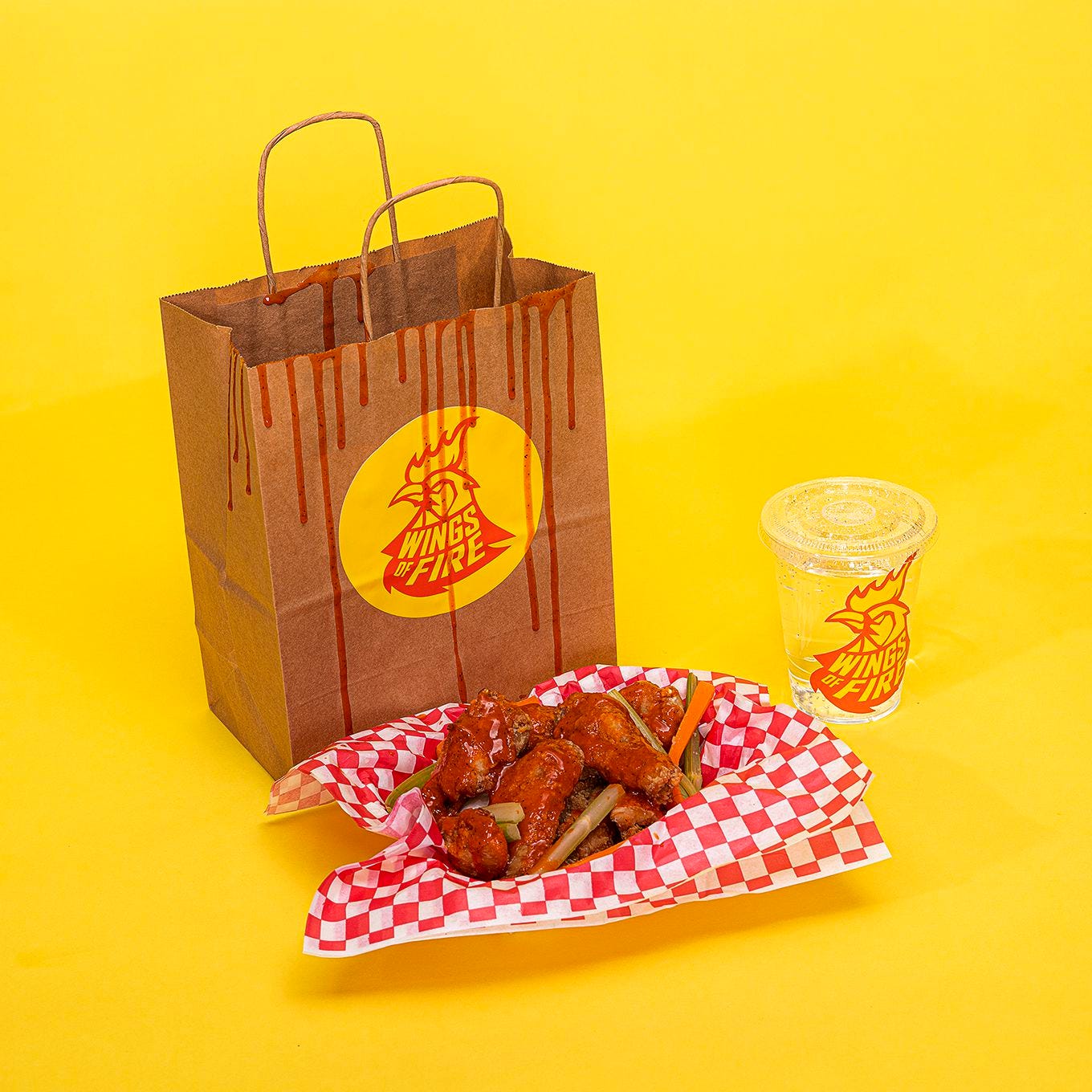 A bag with a sticker for a fake brand called Wings of Fire next to a basket of chicken wings and a clear plastic cup with another sticker for Wings of Fire on a yellow backdrop.