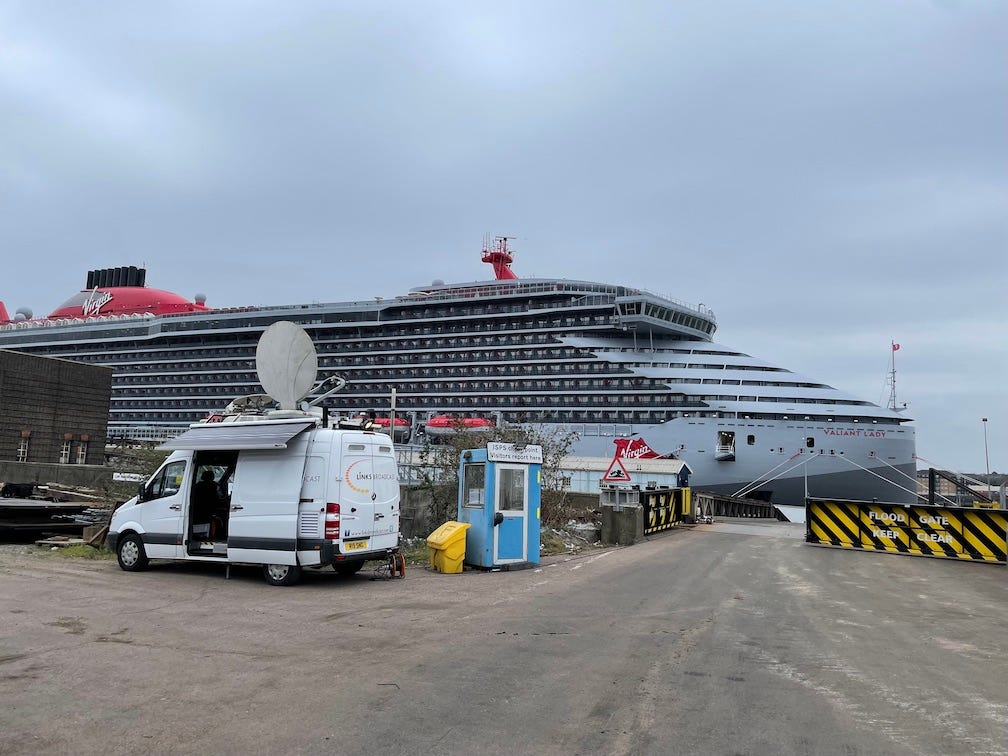 The front 2/3 of a big cruise ship is docked. In the foreground a van with a big satellite dish on top of it in the service area of the dock.