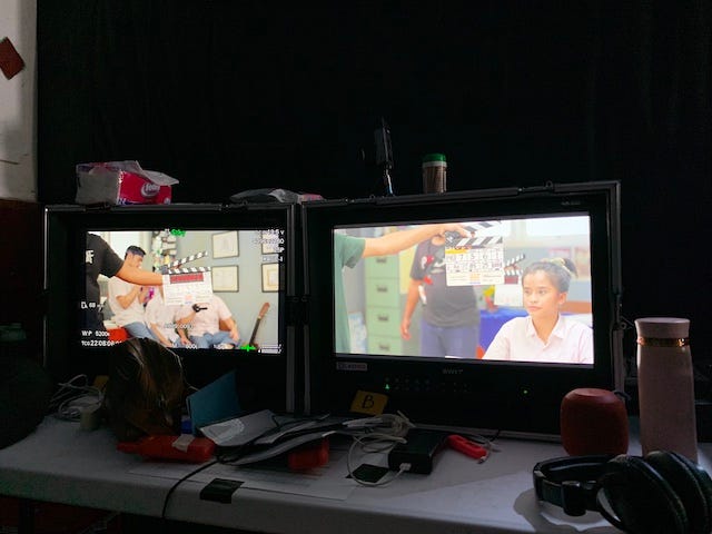Two video monitors showing film clappers and actors in position for a film take. The table is messy with scripts, headphones, tumbler, and other electronics.