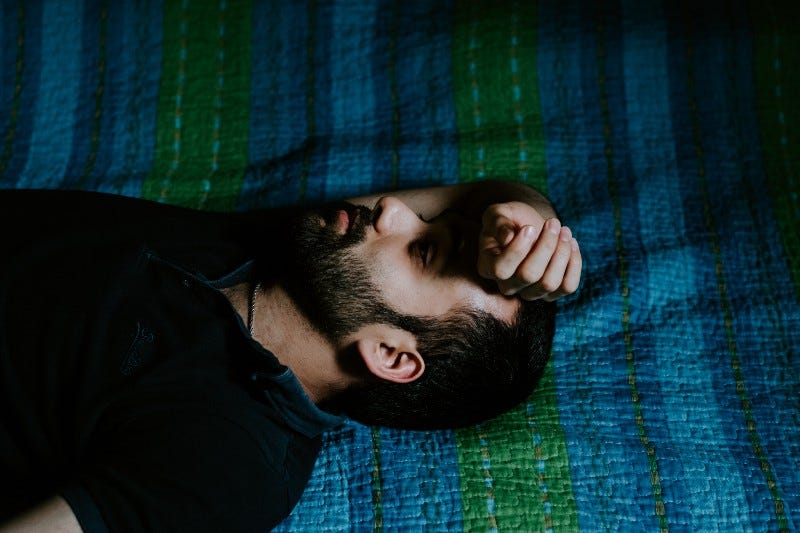 A man lays on a green and blue blanket with his hand placed over his forehead.