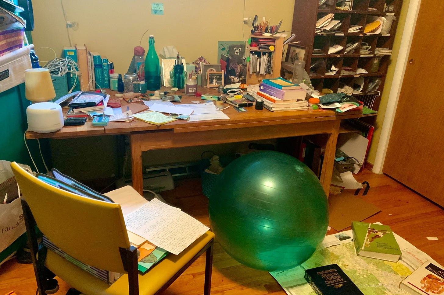 A messy desk with an exercise ball in front of it.