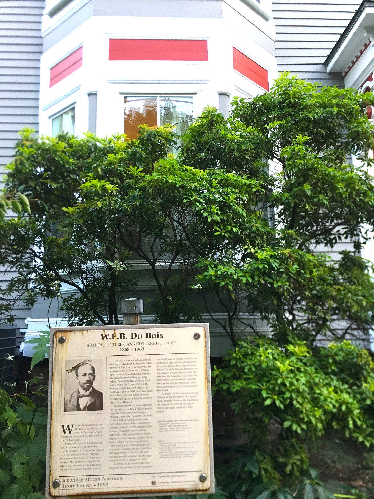 The exterior of the historic W.E.B. Du Bois house in Cambridge, with a commemorative sign backed by trees and shrubs.