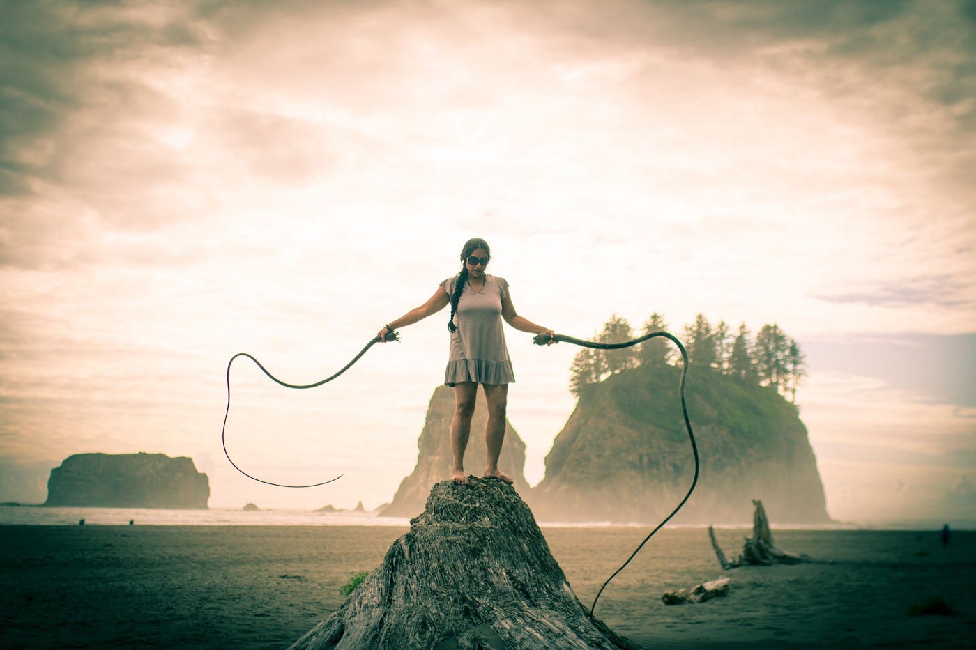 A picture on a beach with the ocean behind and rocks rising from the shoreline. In the center foreground stands a brown-skinned woman on a rock on the beach. She is wearing a grey dress, sunglasses, and her hair is braided. She is holding kelp in each hand and wielding them like whips.