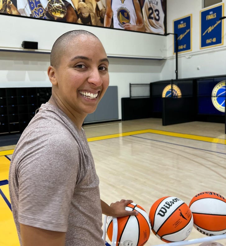 A photo of Layshia Clarendon with their head shaved and a grey-ish shirt on smiling at the camera while standing next to a rack of basketballs on a basketball court that appears to be in the practice facility for the Golden State Warriors. The photo is from Layshia's Instagram.