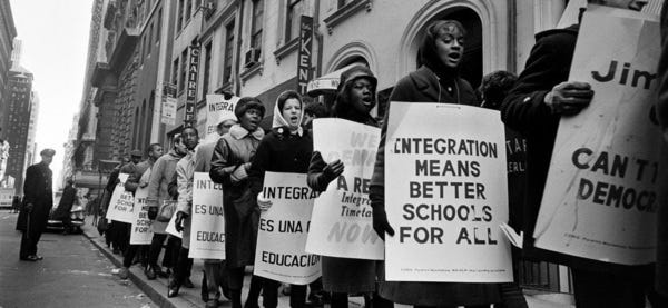 A group of protesters in the civil rights era stand along a street holding signs demanding integrated schools and equality for all.
