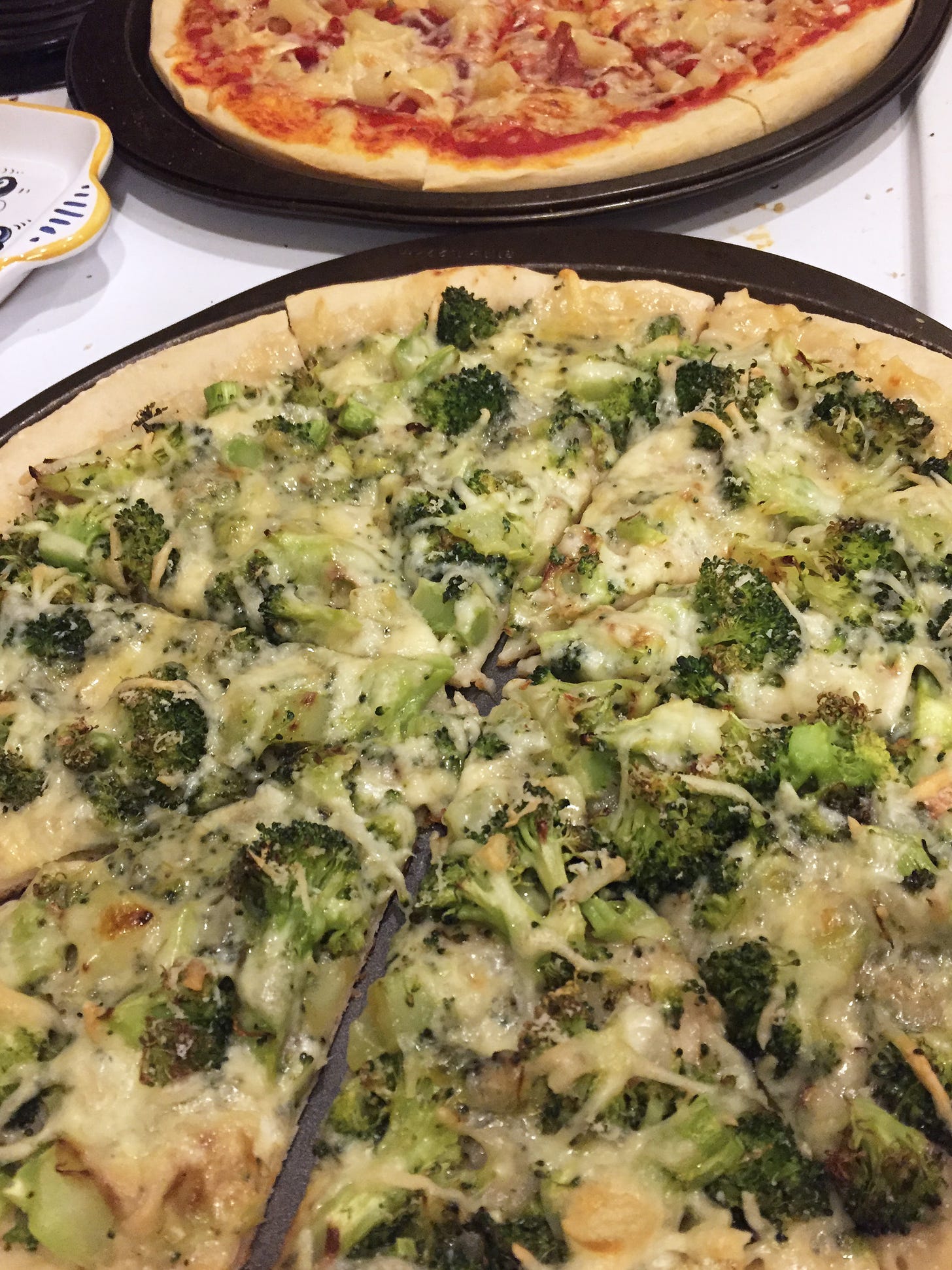 two pizzas in their pans. In the foreground, a broccoli pizza with white sauce. In the background, capocollo and pineapple with red sauce.