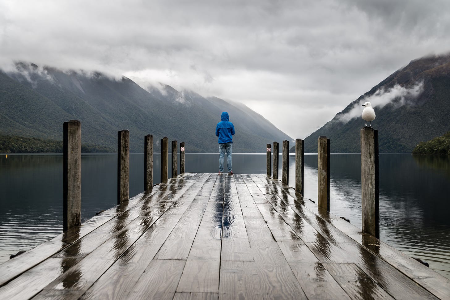 View of a person in a blue hoodie standing at the end of a rain-soaked dock while looking out on a calm lake surrounded by mountains topped in low-hanging gray clouds.