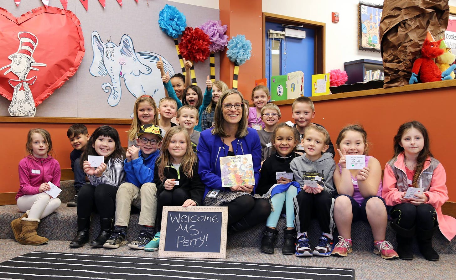 Picture of Superintendent Christy Perry holding a book and siting with elementary students