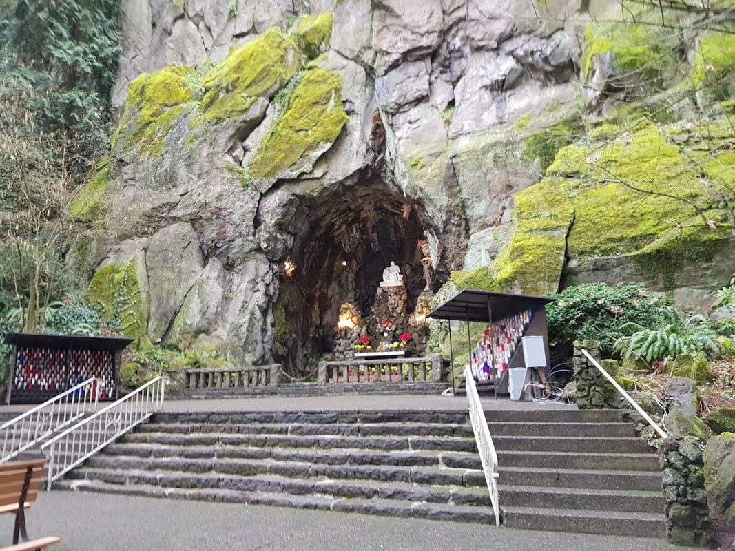 I en liten grotta i en stor klippvägg finns en vit pieta-staty. Framför grottan trappor och ljushållare.