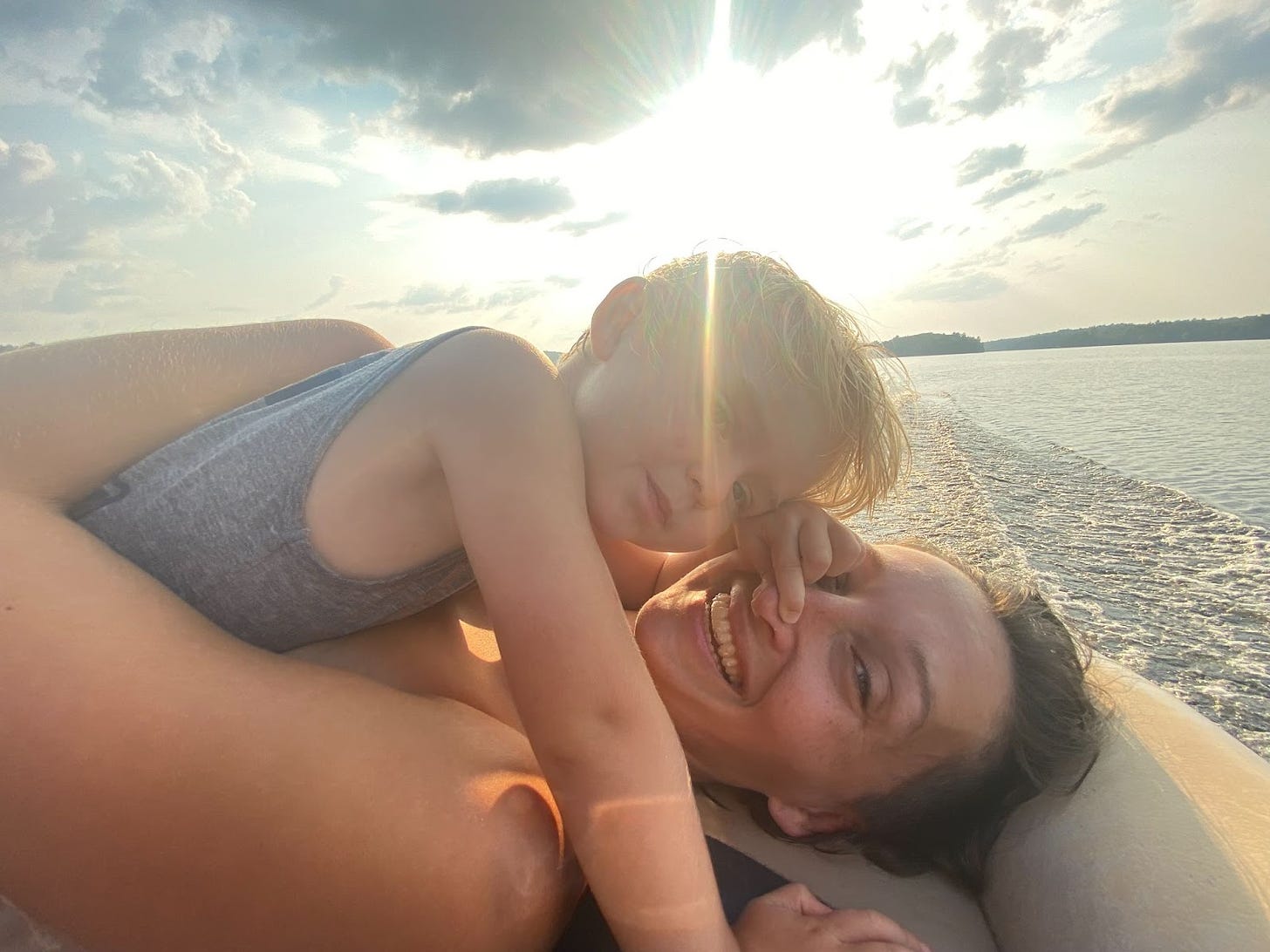 Tarzan laying in the back of a boat with her blond, 3 year old son laying on top of her. The sun sets in the background and you can see it reflecting on the water. 