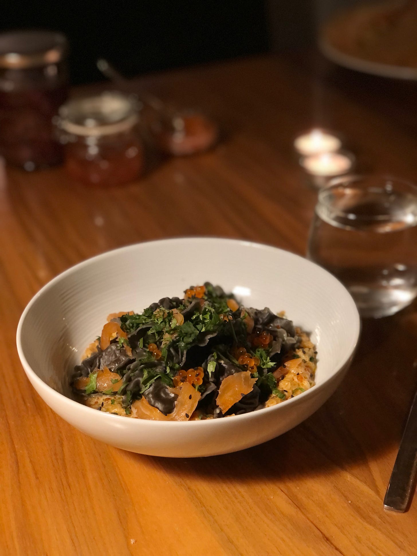 A white poreclain bowl on a wooden table, with a melange unidentifiable bits, some warm browns, some bright orange small spheres, some raggedy black pasta, some coriander. 