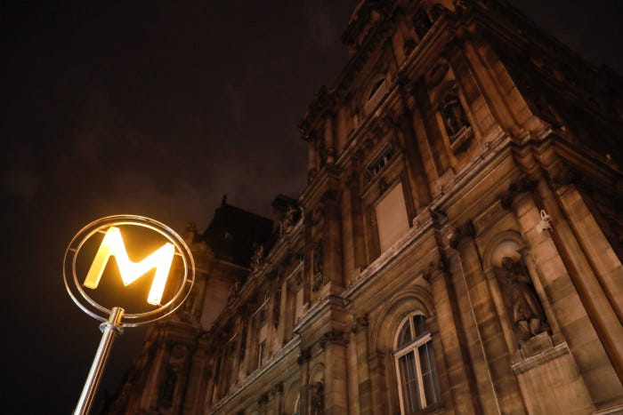 A big ‘M’ is lit up outside a metro station while Paris City Hall is in darkness behind it