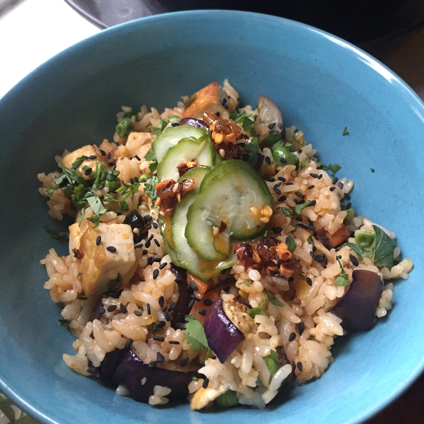 A blue bowl filled with fried rice, with slices of eggplant and cubes of fried tofu visible. On top are thin slices of quick pickled cucumber, black sesame seeds, and chili crisp.