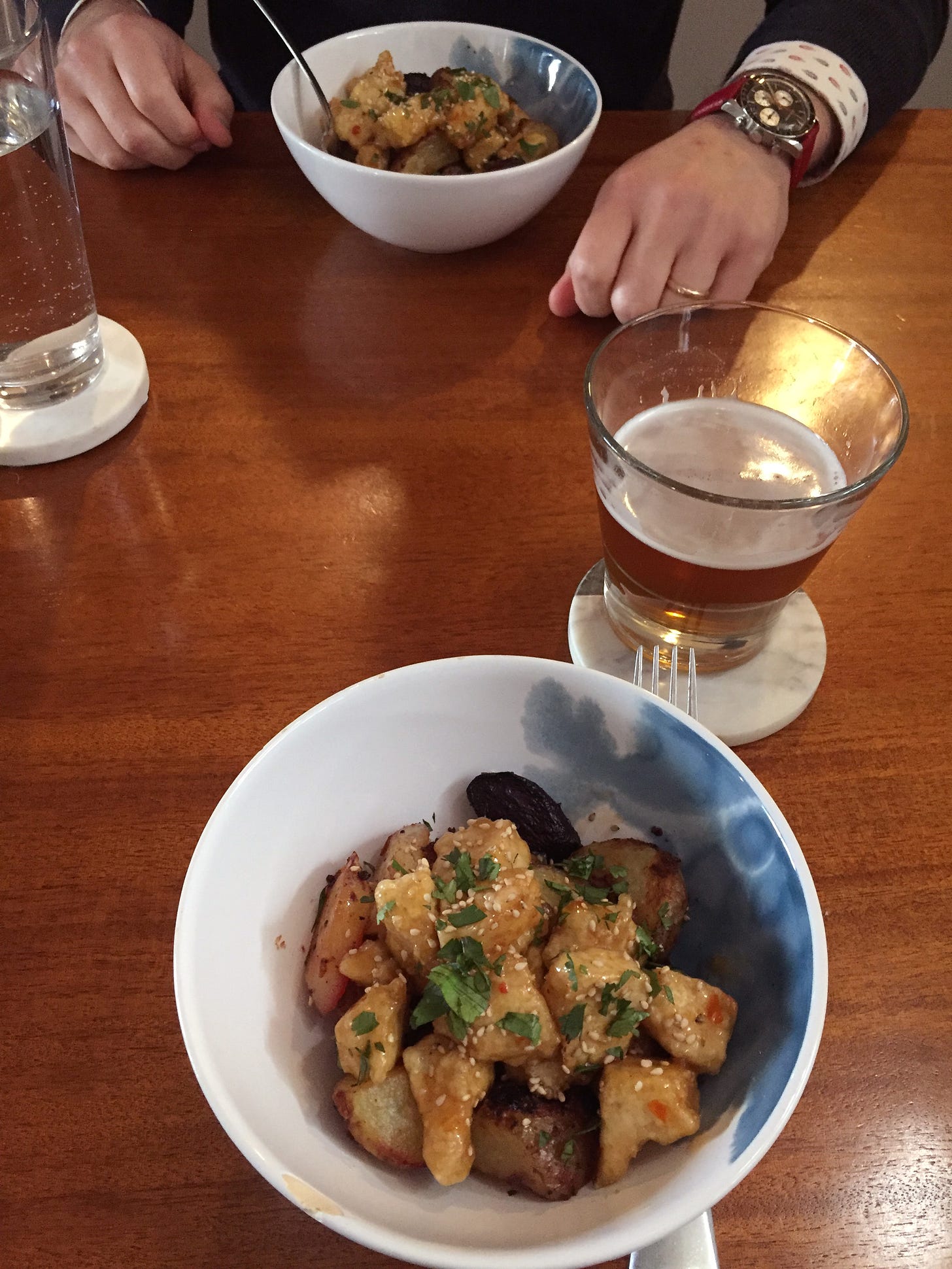 Two white and blue bowls on the table across from each other, and a beer and a glass of water on coasters. In the bowls are pieces of the crispy tofu coated in sweet chili sauce and sprinkled with sesame seeds and cilantro. Just visible below the tofu are pieces of potato, purple carrot, and beet.