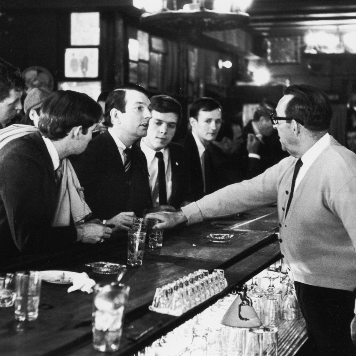 A group of white men in suits stare down a white bartender putting his hand over the glass of a beer he's just served them.