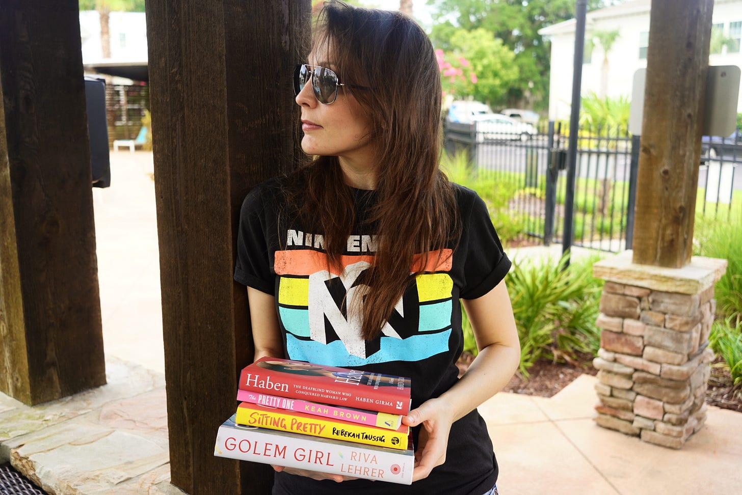 a photo of Kendra, a 30-something white woman wearing sunglasses and black Nintendo t-shirt, holding a stack of books, including The Pretty One, Golem Girl, Haben, and Sitting Pretty.