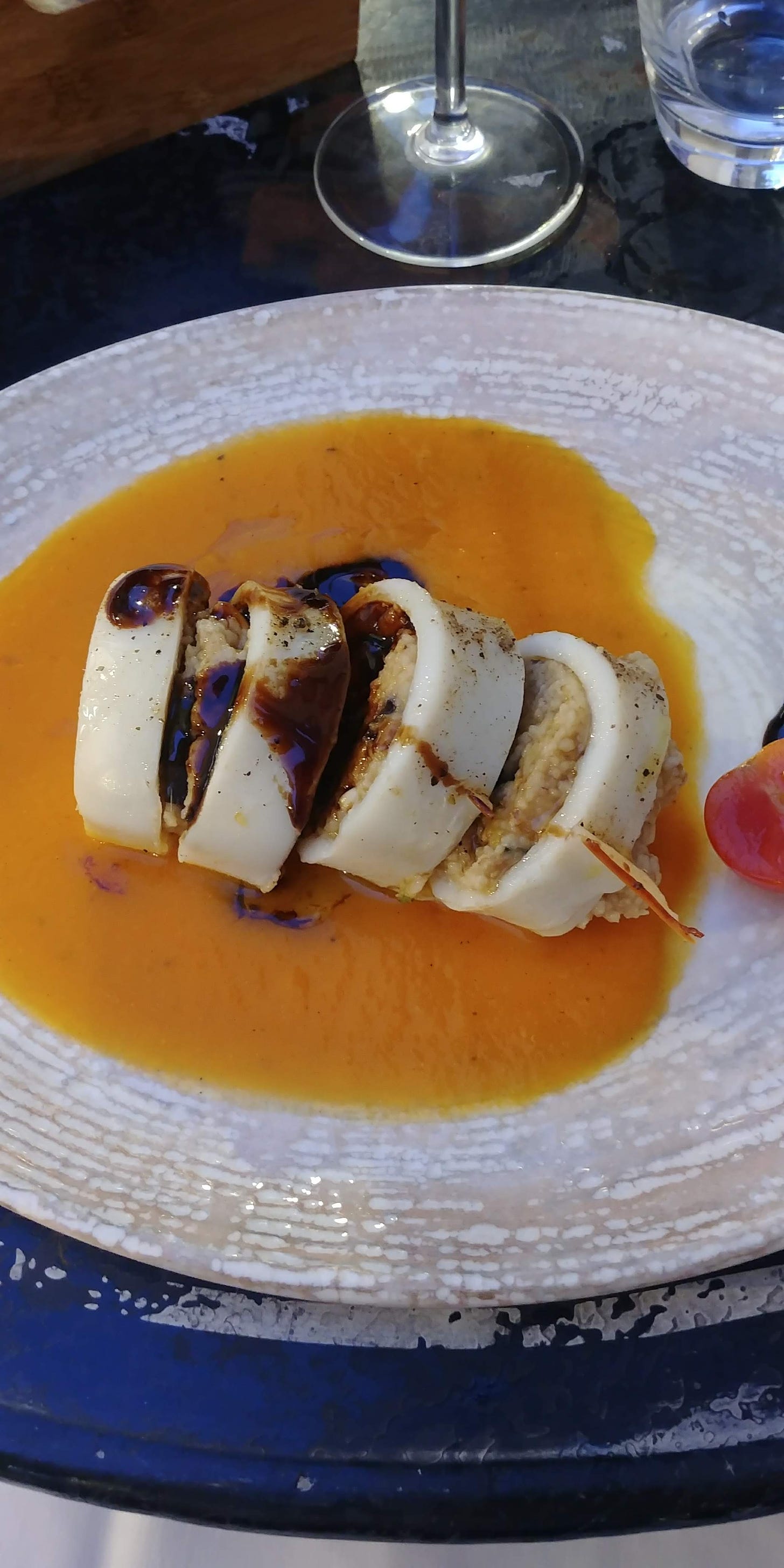 a photo of stuffed squid with couscous. Grainy couscous fills white rounds of squid flesh, cut into slices, on an orange sauce. The dish is served on a round white plate. The base of a wine glass is visible at the top of the image 