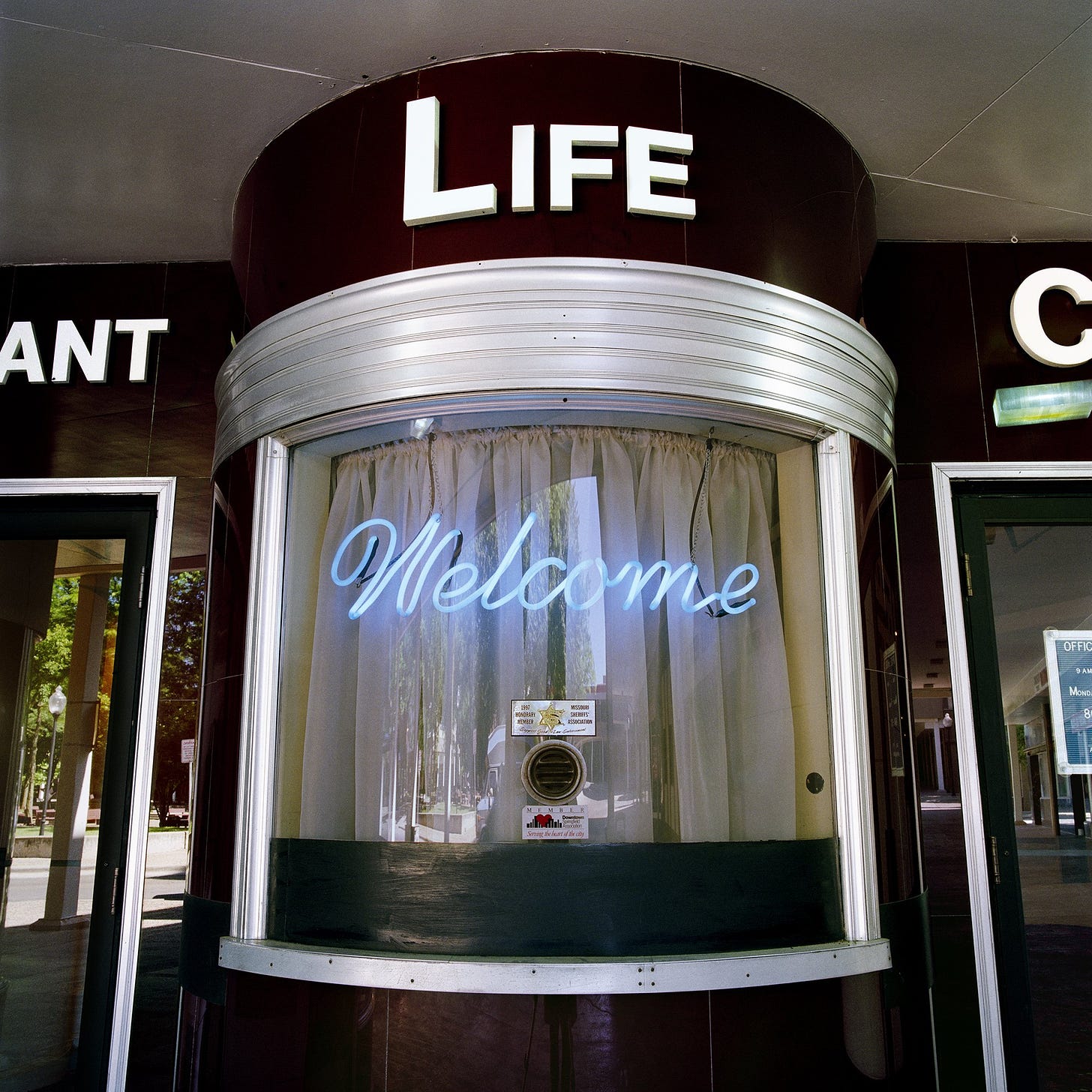 a ticket window in a mid-century movie theatre with the word "Welcome" rendered in blue neon and hanging in a curved window