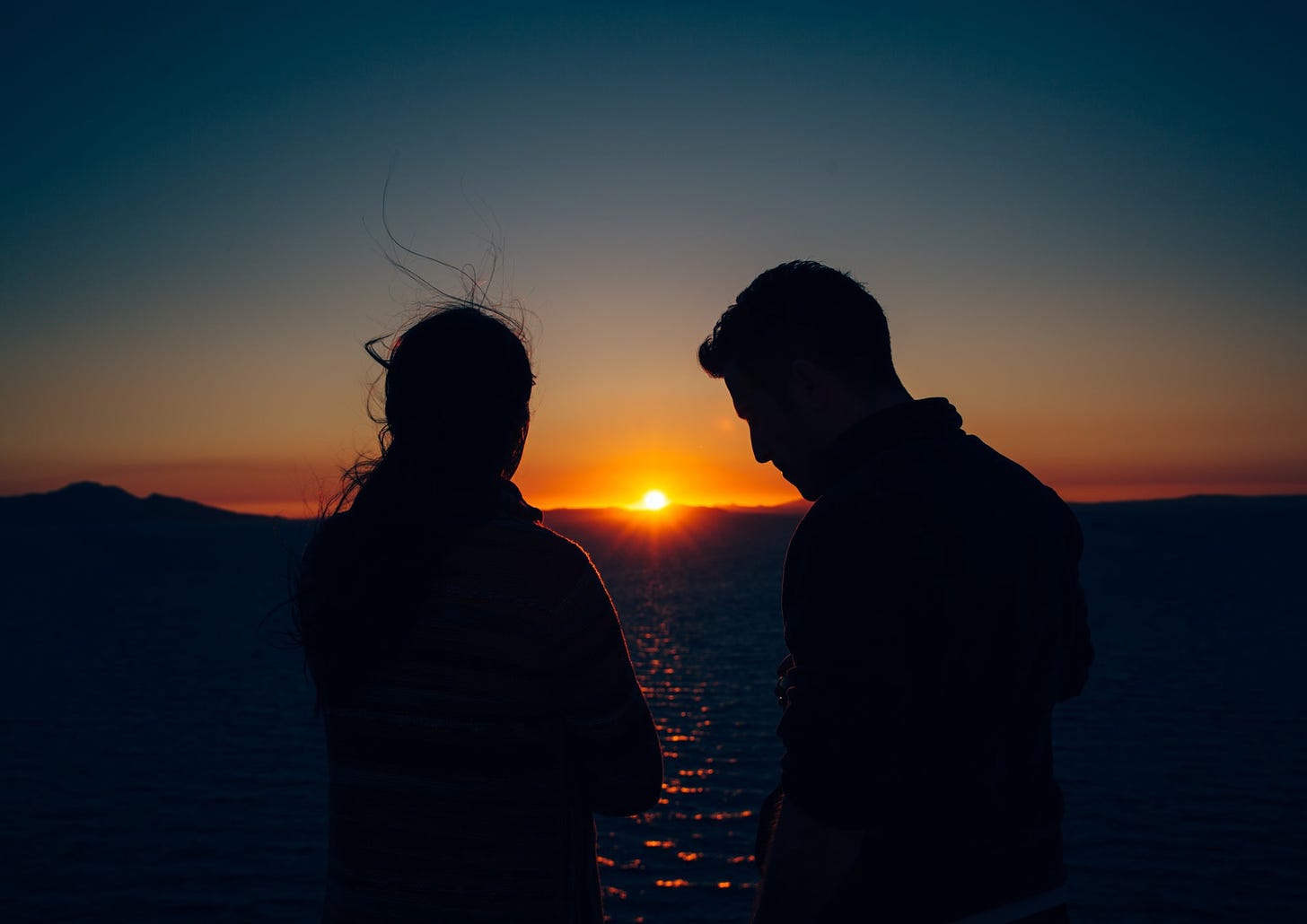 A couple stands apart as the sun sets over a beach horizon.
